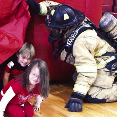 Taylynn Stafford follows Knyx Scoggin to escape the simulated burning house while firefighter Alex Tyler holds the flap open for Amber Hansen’s first-grade class in the Nance Elementary Gym during the Clinton Fire Department’s presentation. CDN | Cale Fire safety education
