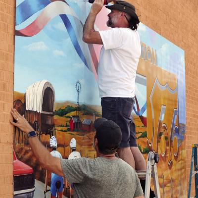 Patrick Hanscom, front, and Scott Henderson install the “American Thread” mural, celebrating the centennial celebration of Route 66 as well as the 250th anniversary of America, on the west wall of S&amp;D Drug. CDN | Christian Jacobsen